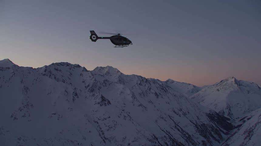 Helicopter flies over narrow valley between snow covered mountains under red tinted sky