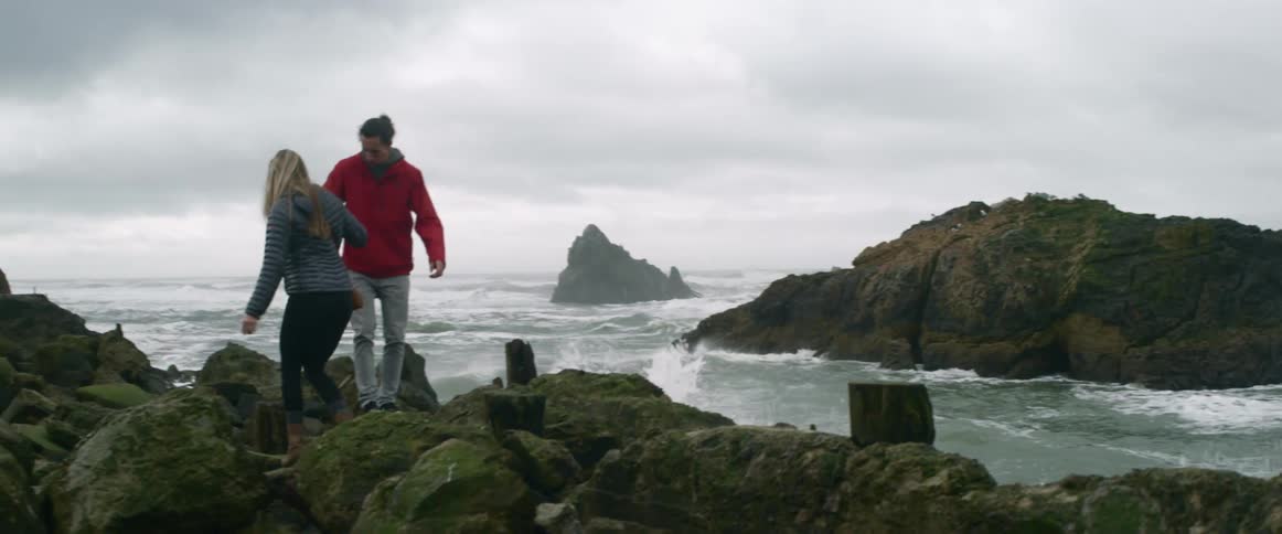 A man helps a woman climb along a rock jetty on the beach