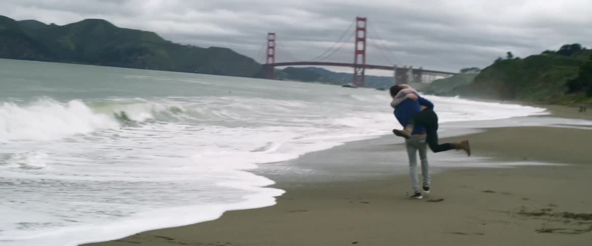 A couple run from waves before the man picks them woman up and playfully heads back towards the water