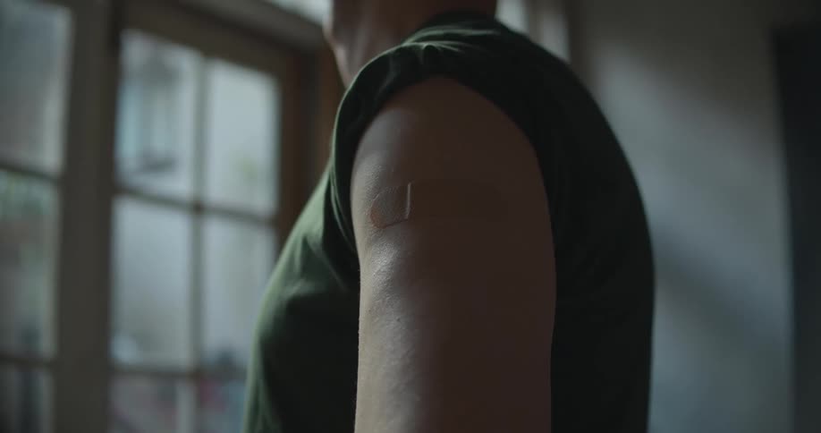 A man stands with his shirt sleeve rolled up with a bandage on his upper arm
