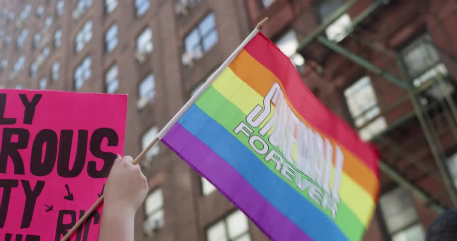 Protesters marching with signs and a rainbow flag on a city street