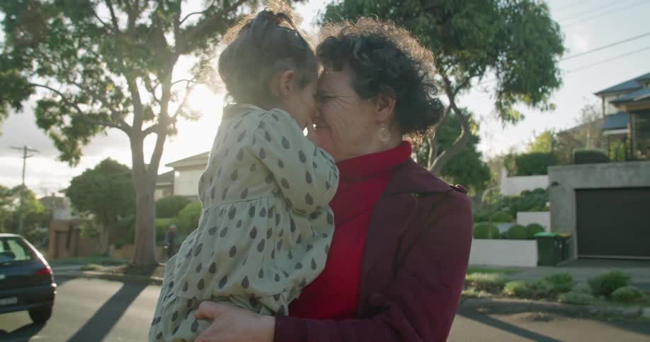Grandmother holds granddaughter in her arms and they rub noses with a smile standing near street in residential neighborhood