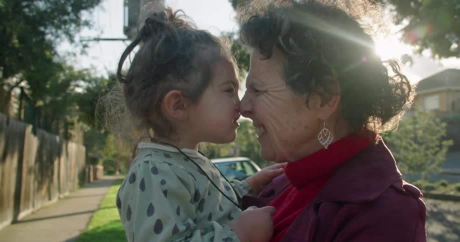 Grandmother holds granddaughter smiling and laughing and giving rubbing noses together standing near street in neighborhood on sunny day