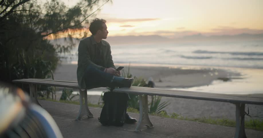 Man sits on a bench near the beach and looks at his cellphone as a pedestrian walks by