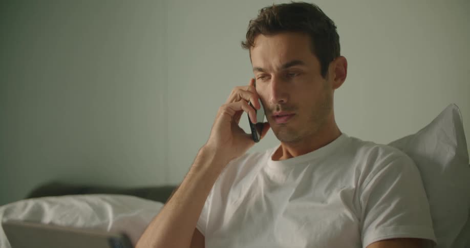 A man types on his laptop with one hand while talking on the phone in bed