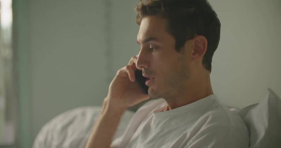A man in the bedroom sitting on a bed talking on a smart phone and using the laptop
