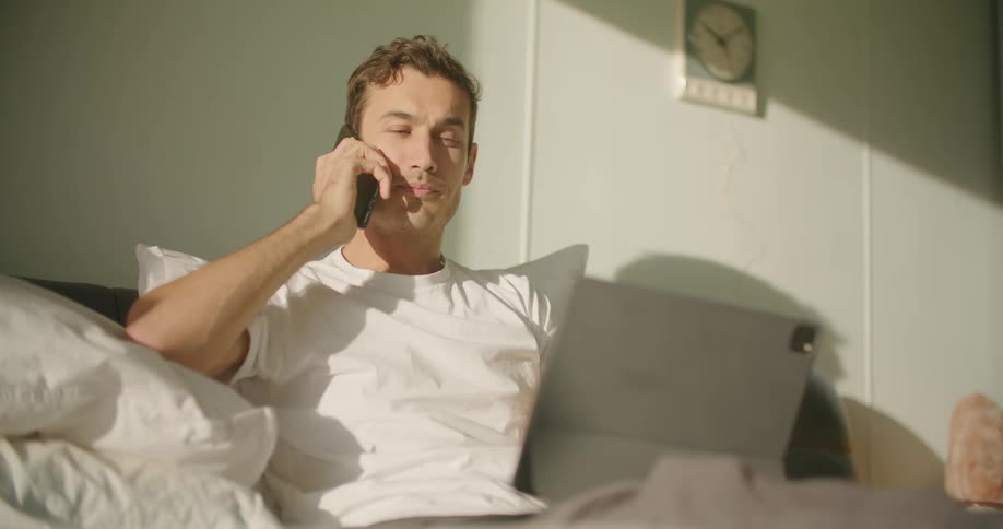 Man sitting in bed talking on the phone while looking at a tablet