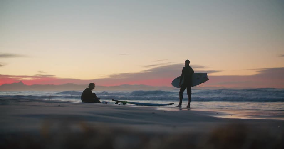 The silhouette of two surfers on the beach looking out at the waves at sunset