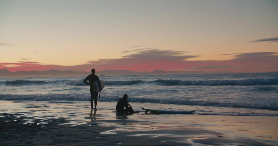 Two surfers sit by the shore at sunset