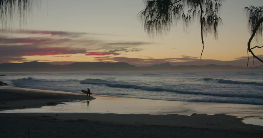 The silhouette of a surfer carrying his board and sitting down on the wet sand watching the waves at sunset