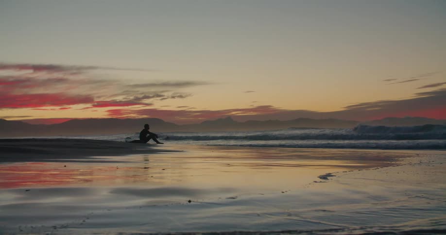 A surfer sits on the beach and watches the sun set as waves crash around him