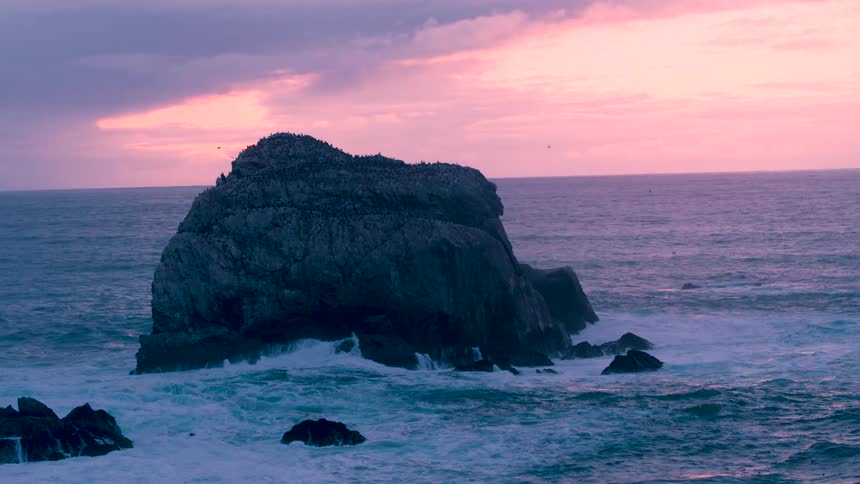 Many birds sit on a big rock in the middle of the sea while the waves crash into the rock