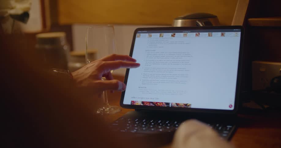 A man's hand taps and scrolls on the touch screen of a laptop as he looks at a recipe with a glass of wine beside him