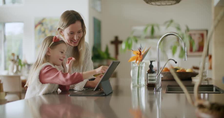 A woman laughs and smiles while watching a young girl using a touchscreen laptop computer on a kitchen counter