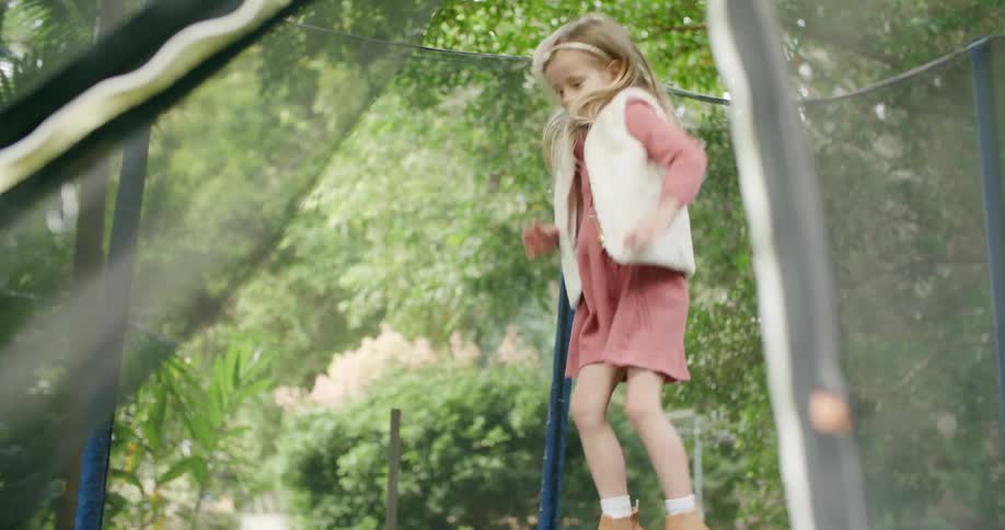 Blonde girl wearing dress and vest jumps on trampoline surrounded by protective netting that is not completely fastened