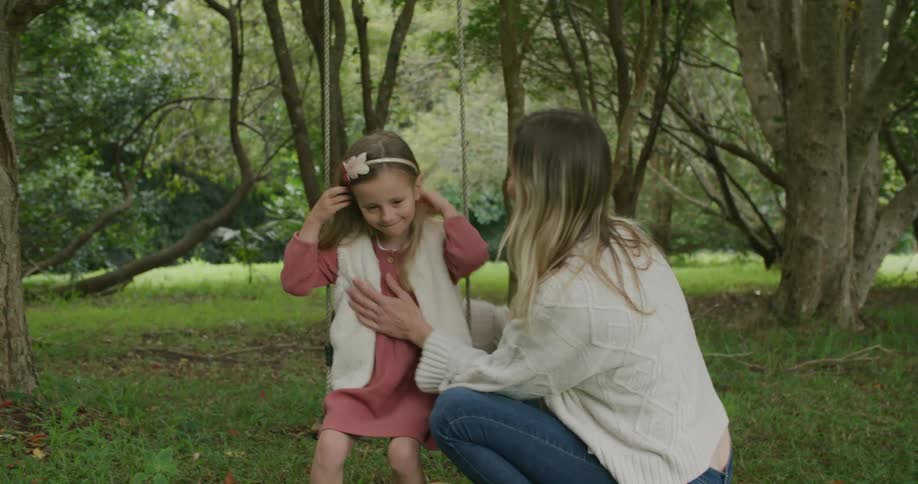 A young girl sits on a swing outside and a woman brushes the hair away from her face