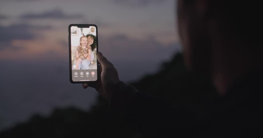 A man waves back at his phone screen where his wife and daughter are waving at him