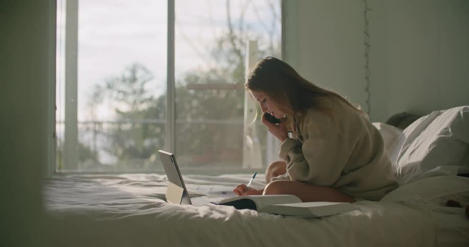 Woman sitting in bed talking on her phone working in front of a tablet