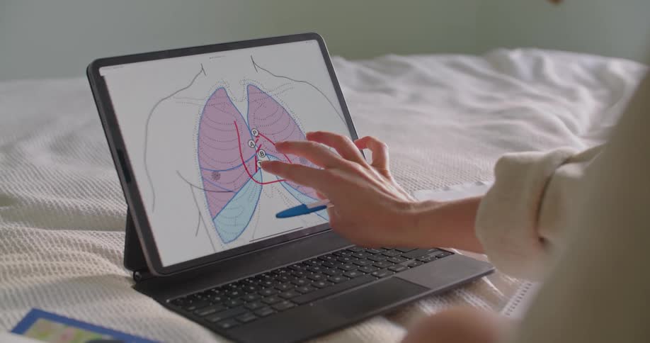 A woman sitting cross legged on a bed uses her fingers to enlarge a lung diagram on her laptop