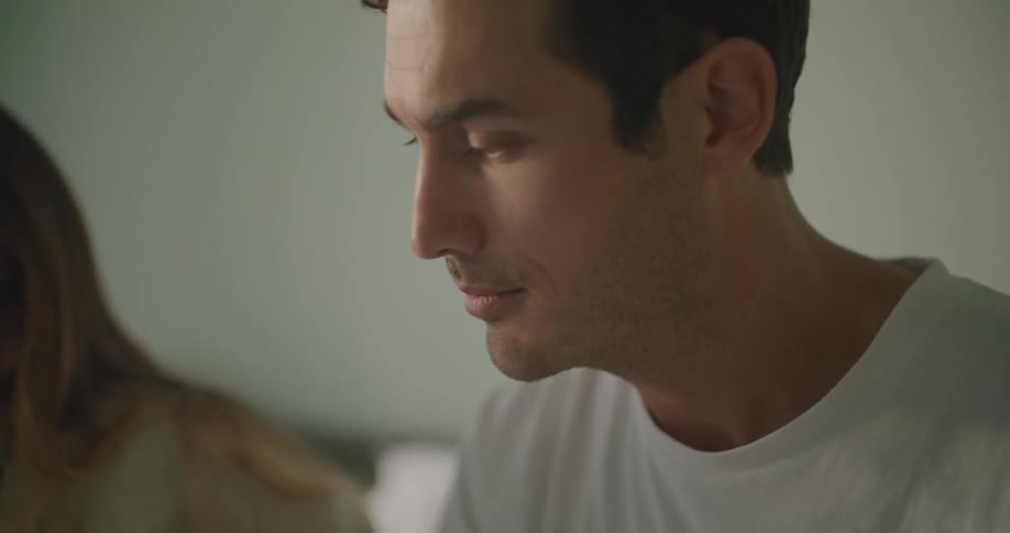 A man touches the screen of a laptop and speaks while sitting next to a woman on a bed