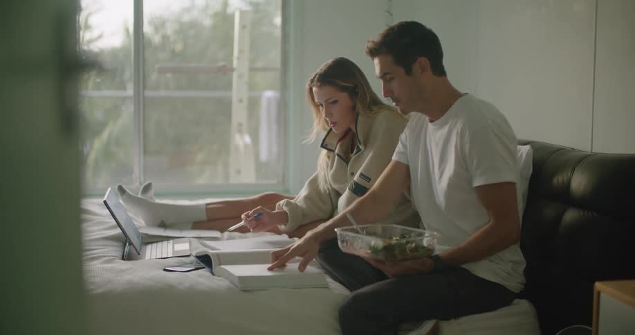 A man and woman sit on a bed together with a large book and laptop