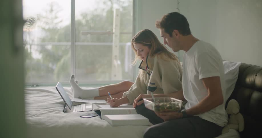 A man and woman sit on a bed looking at a laptop and writing in a large book