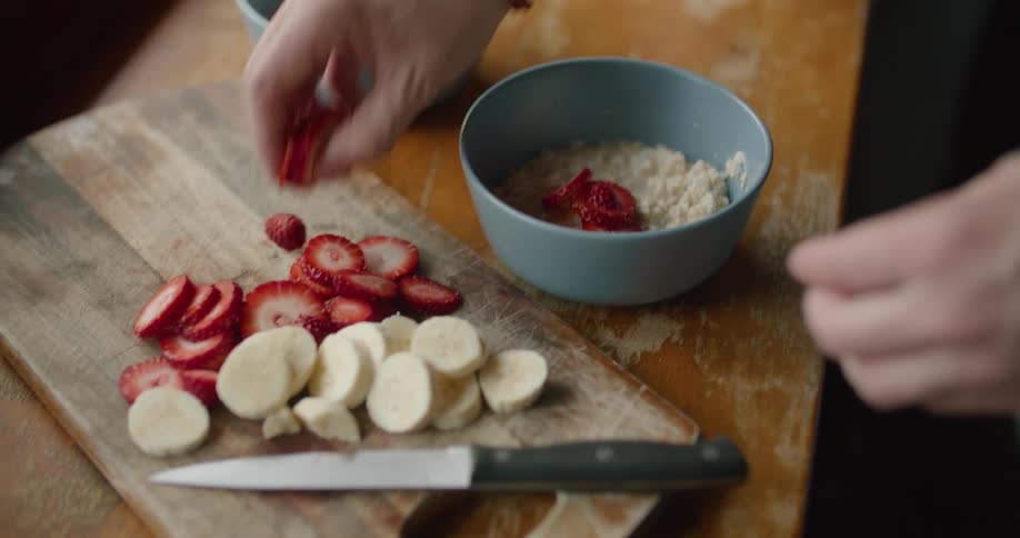 A person preparing bowls of oatmeal with sliced strawberry and bananas