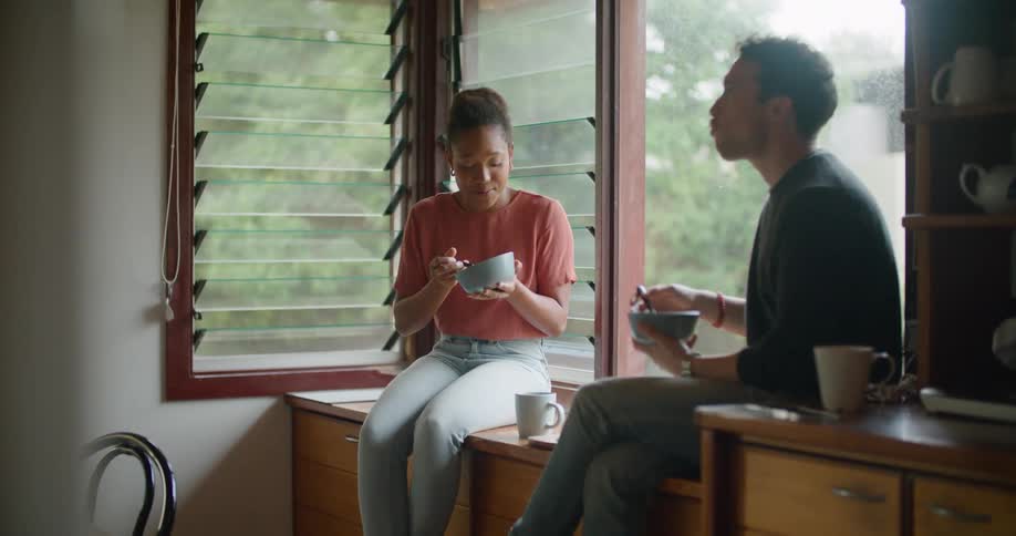 A man and woman sit by a window in a house and eat and talk