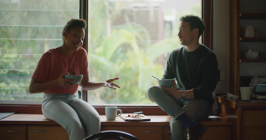 Man and woman sit holding food bowls near window showing trees and another building as they gesture and talk
