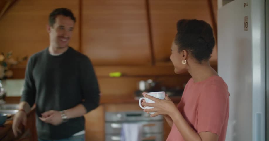 A woman holding a mug stands in a kitchen talking and smiling with a man