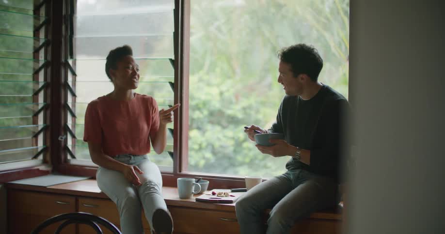 A man and a woman in a room sitting and talking with a wall and windows in the background