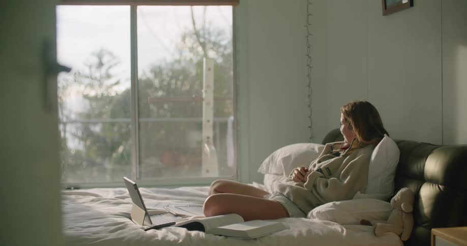A woman sitting on a bed with a laptop with a door and wall in the background