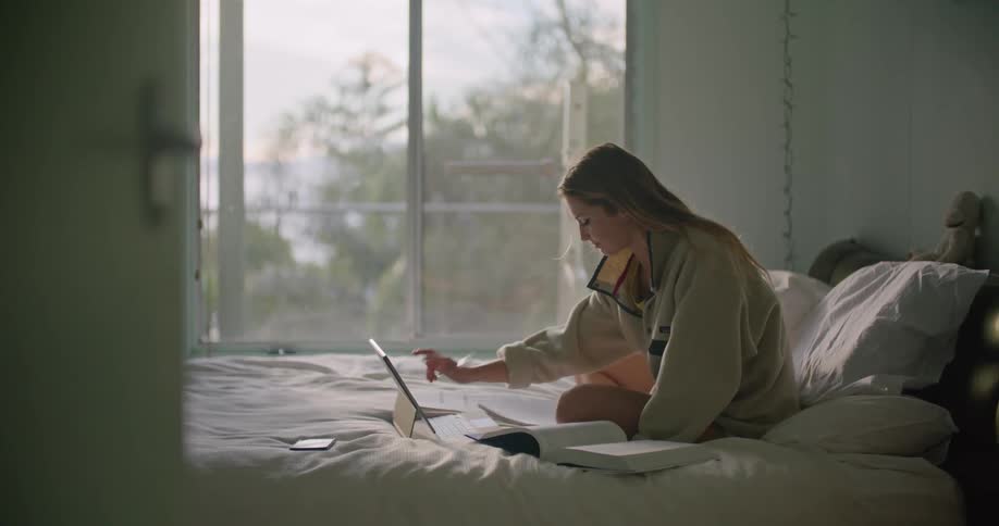 Blonde woman wit oversized sweatshirt sitting on bed writes in notebook while using tablet