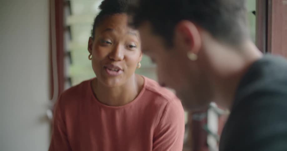A man and woman gesture at the screen of a tablet as they talk while sitting next to a window