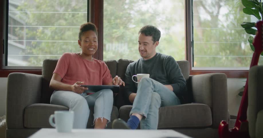 Young couple sitting in the couch at home talking and smiling while looking at a tablet