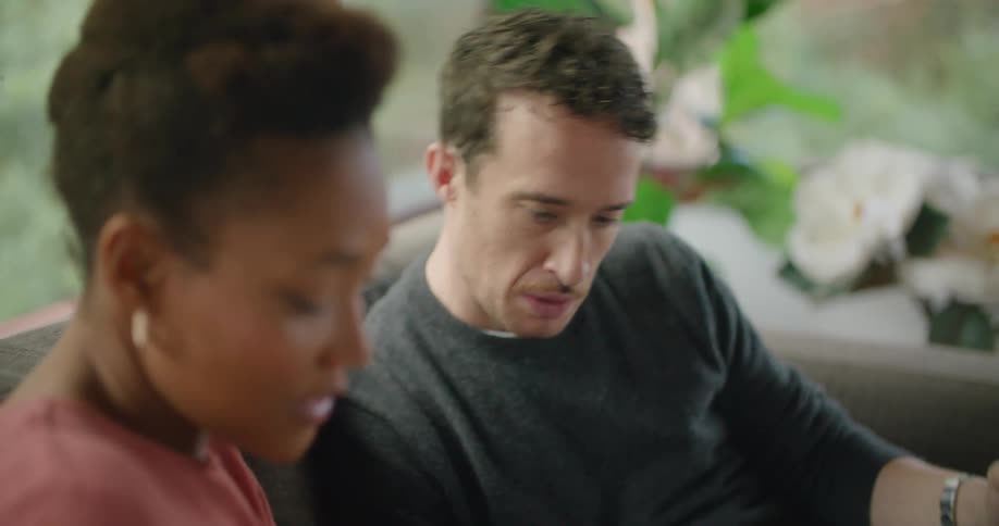 A man and a woman in a room sitting on a couch looking at a tablet