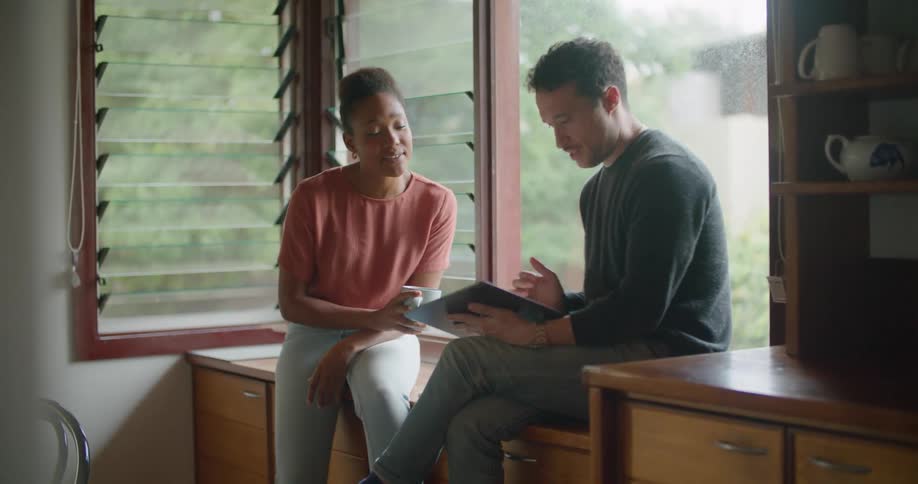 Two friends sitting in the countertop of the kitchen talking while using a tablet