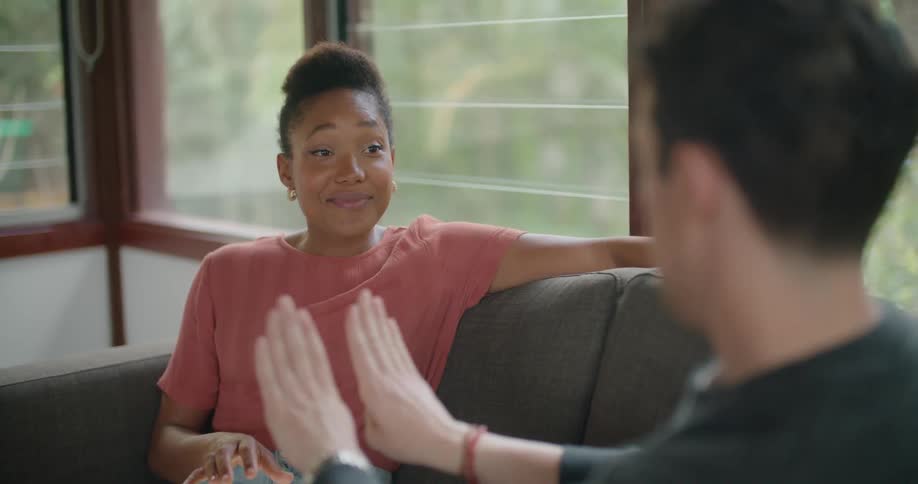 A man and a woman in a room sitting on a couch and talking with a wall and window in the background