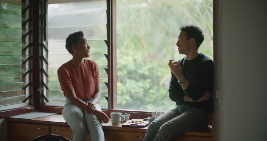 A man and a woman in a room sitting and talking with a wall and windows in the background