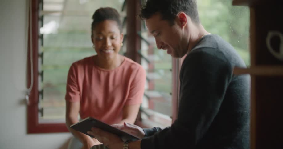 Man wearing grey sweater and jeans uses tablet next to woman with short hair red shirt on window pane