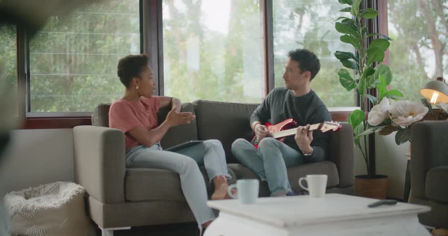 Man plays guitar sitting on couch next to woman as they talk