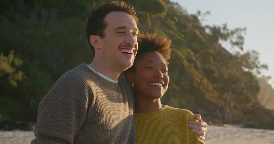 Man with grey sweater holds woman with short hair and yellow sweater on the beach at sunset