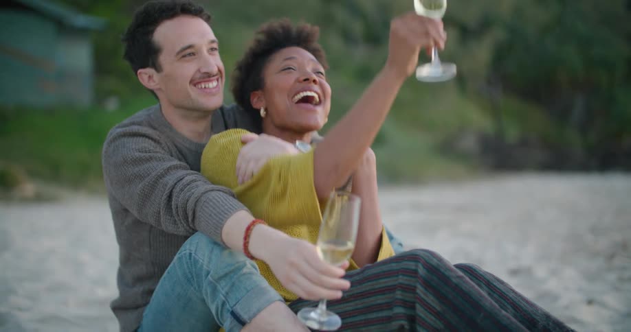 A man and woman sit on the sand embracing laughing and holding champagne flutes