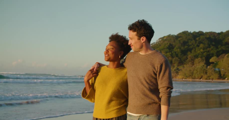 A man and woman speak while walking along a beach with their arms around each other during the day
