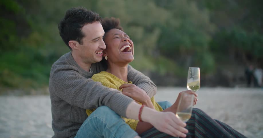 A man and woman sit on a beach together with glasses of champagne