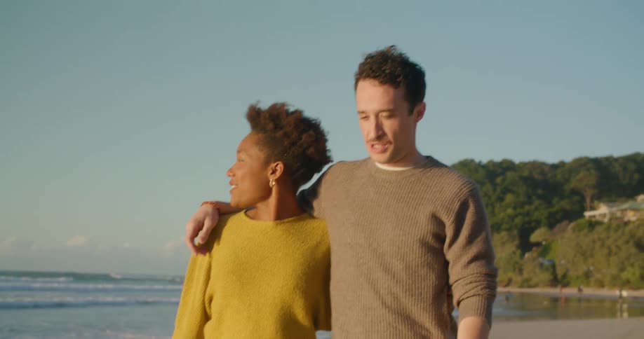 Two people talk as they walk arm in arm down the beach