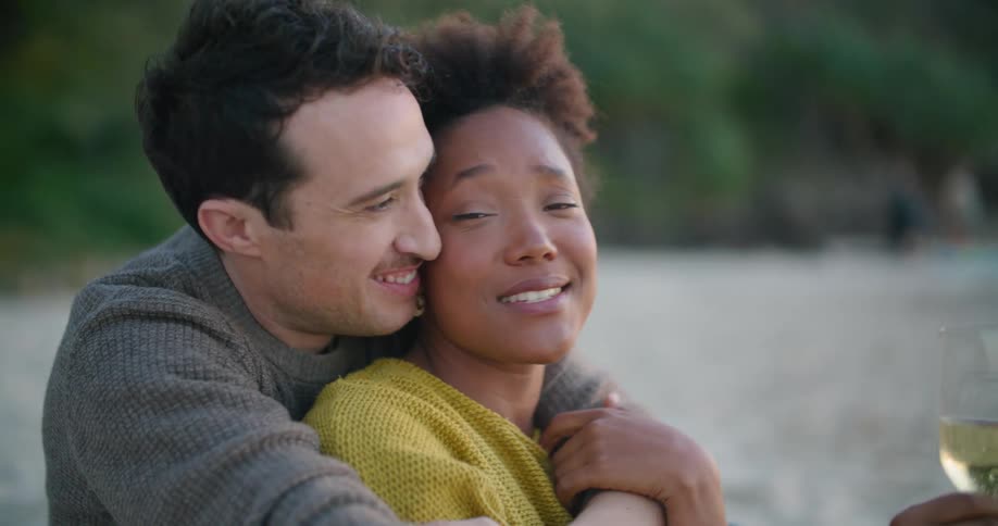 A man hugs and kisses his girlfriend as they drink wine on the beach