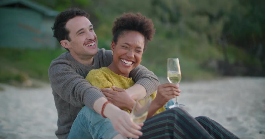 A man sitting on the sand hugs a woman as they laugh and talk both holding champagne flutes