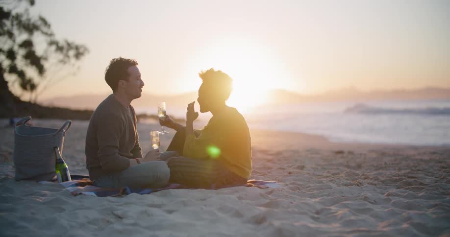 A woman leans back laughing before clinking champagne glasses with her man on the beach