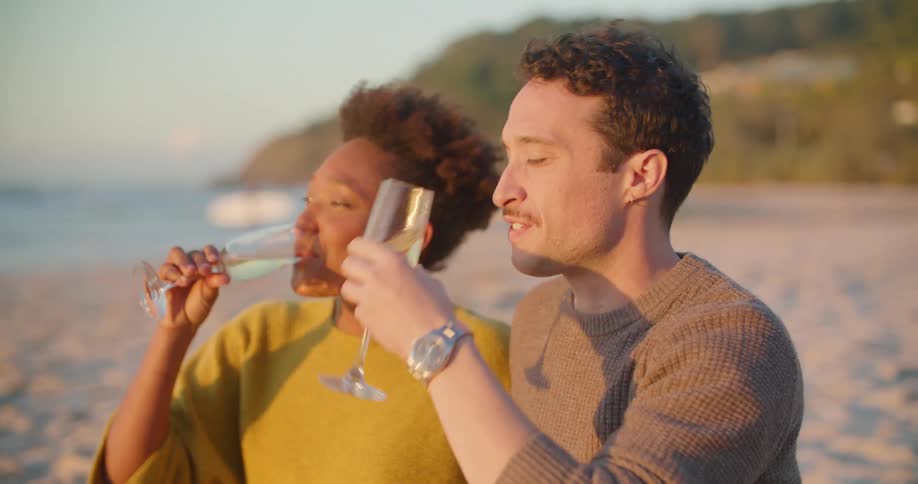 A man and woman sit on the beach and laugh and talk as they drink champagne